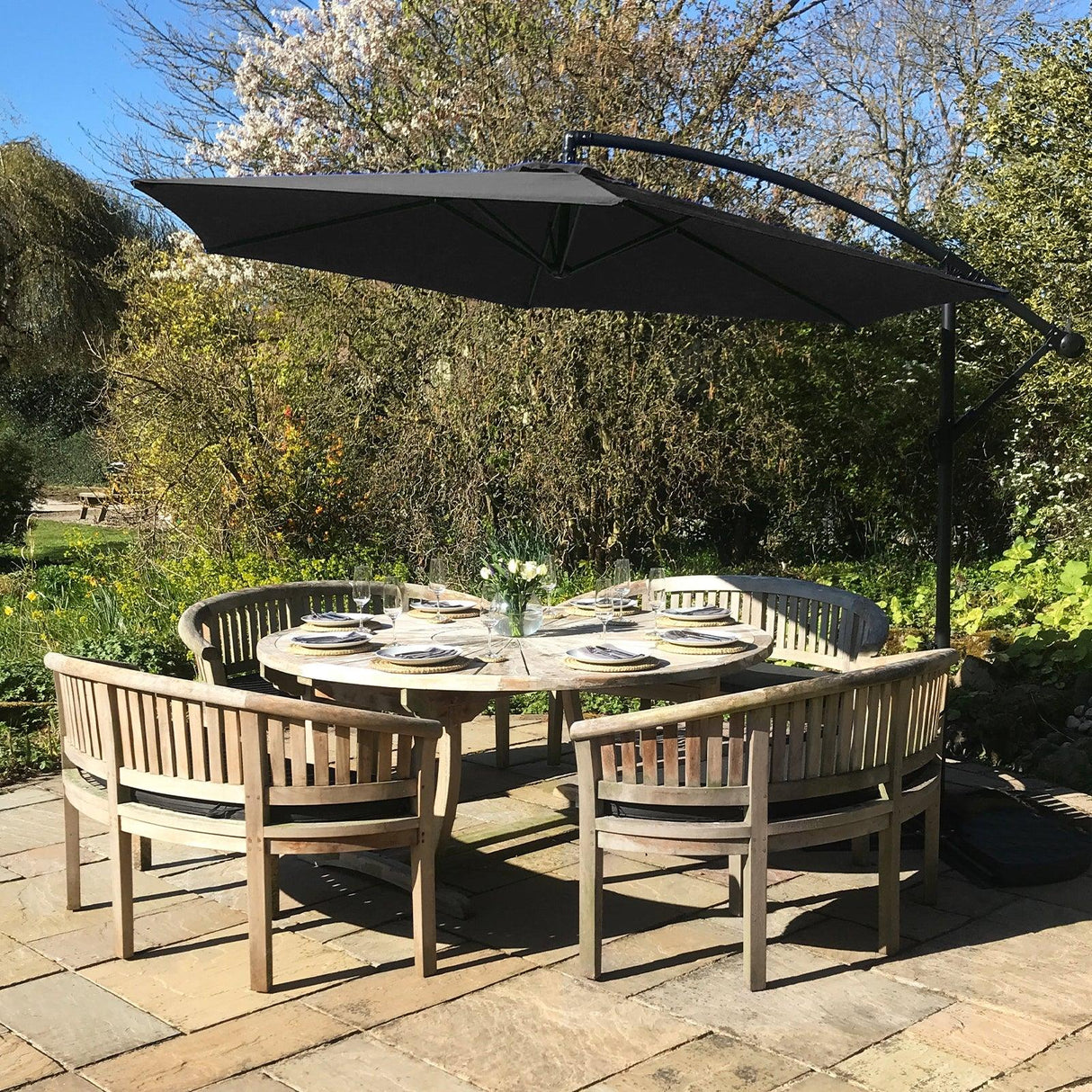 Black cantilever parasol with a sleek black canopy, providing shade over a round wooden dining table surrounded by curved benches in a garden setting