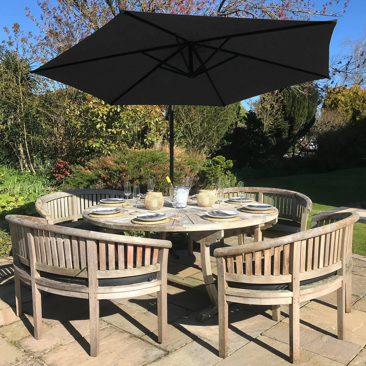 Black cantilever parasol providing shade over a round wooden dining table set with plates and glasses in a garden setting