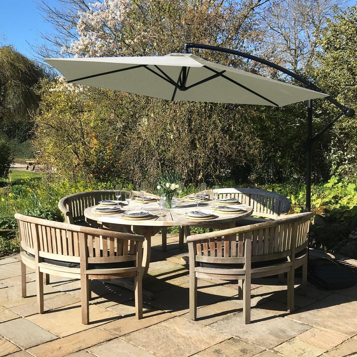 Cream cantilever parasol in a garden setting, featuring a large beige canopy, curved black frame, and surrounded by wooden dining chairs and table