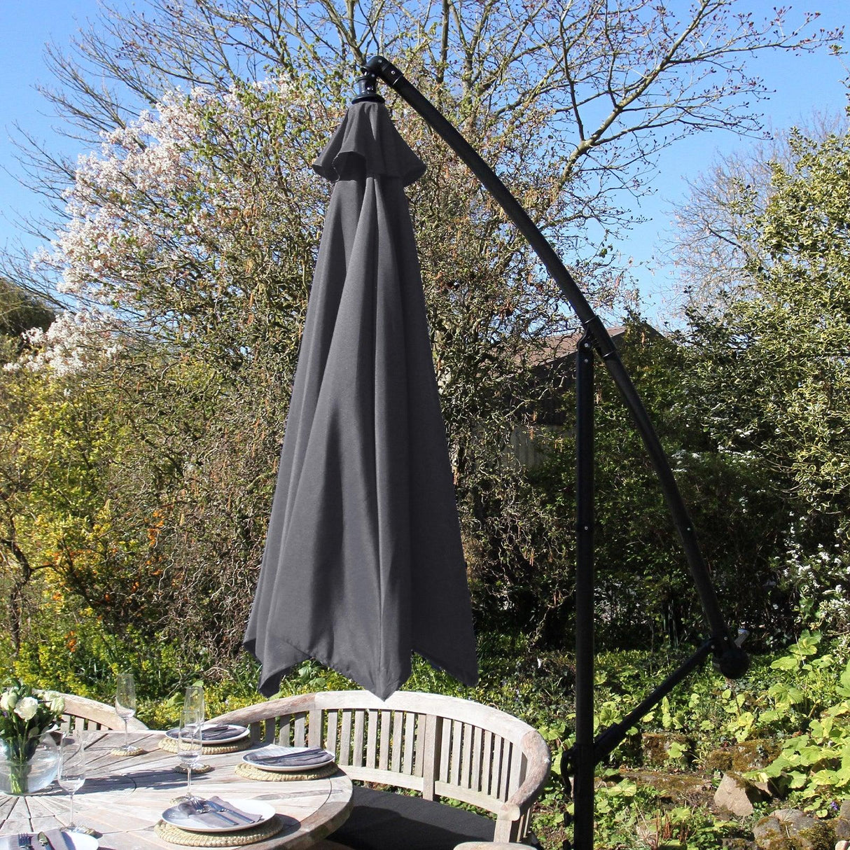 Grey cantilever parasol with a sleek, curved arm and a large canopy, set in a garden with blooming trees and outdoor dining furniture