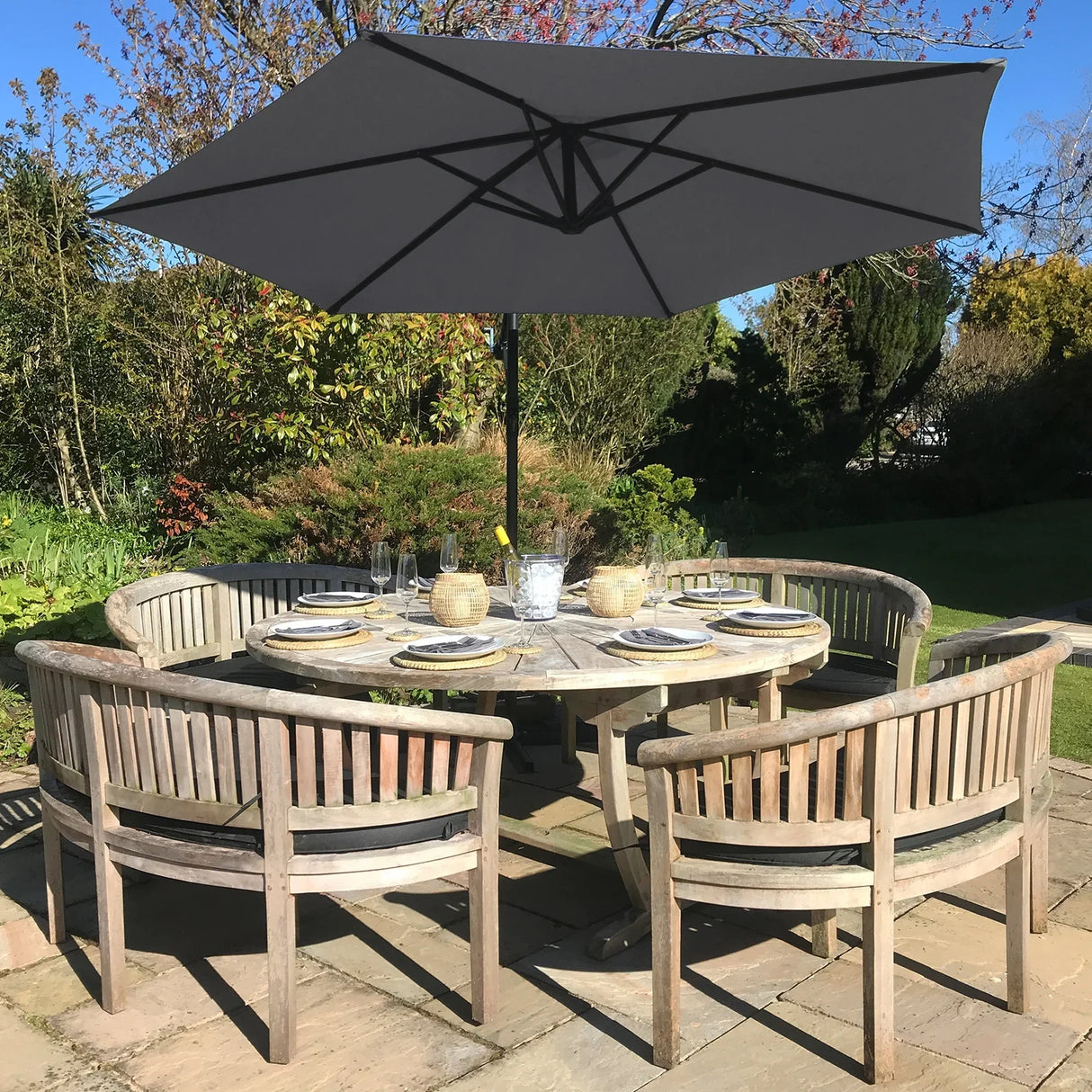 Grey cantilever parasol with a square base, providing shade over a round wooden dining table set with plates and glasses in a garden setting