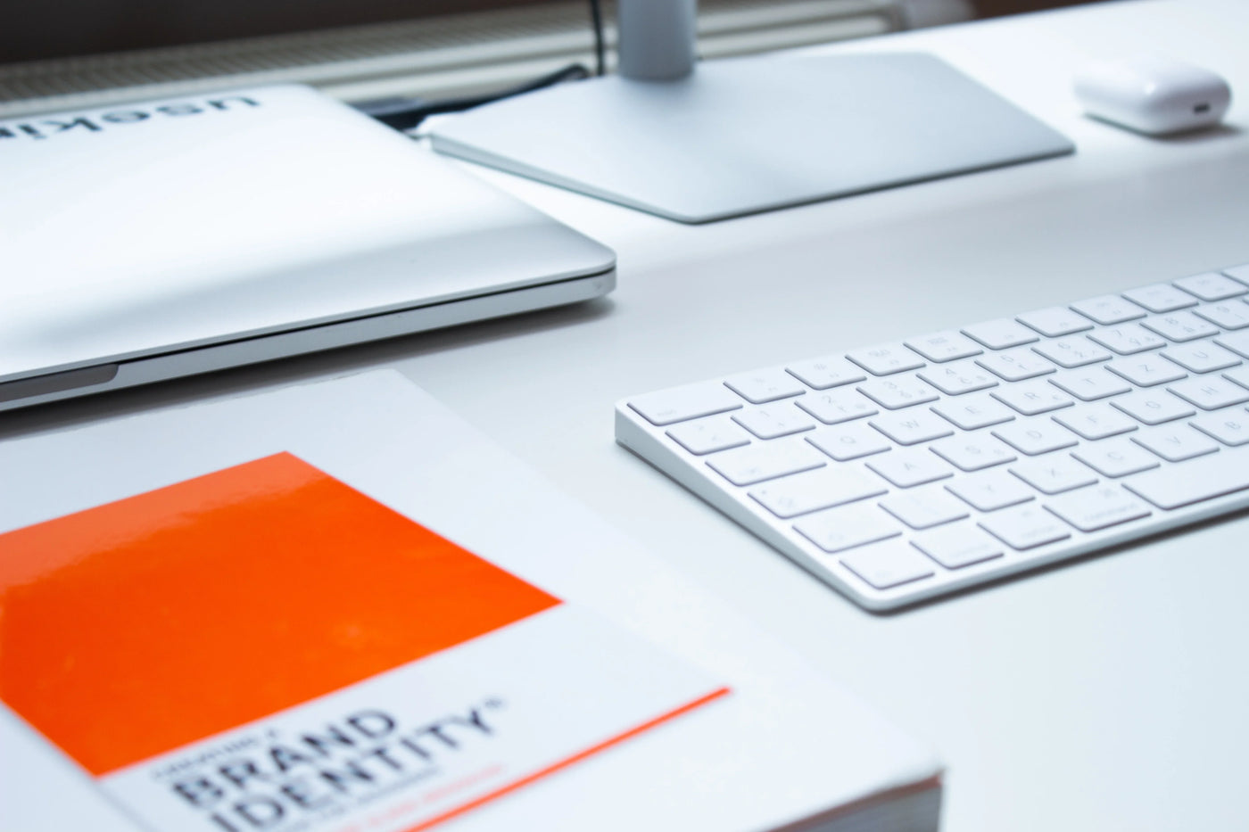 A sleek workspace featuring a silver laptop, white keyboard, and an orange-covered book titled "Brand Identity" on a clean desk