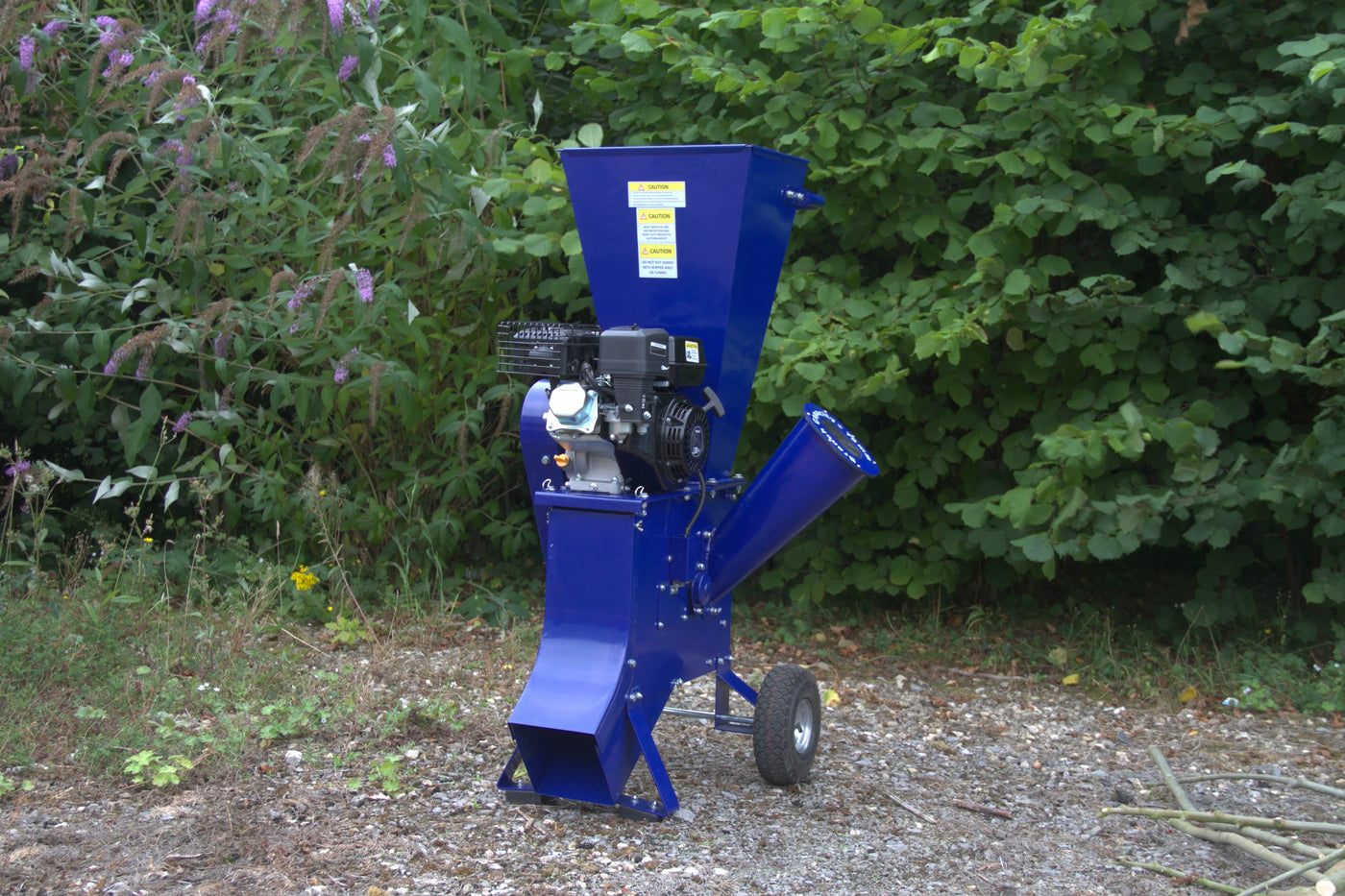 Blue wood chipper with a powerful engine, featuring a large hopper, side chute, and sturdy wheels, set against a green foliage backdrop