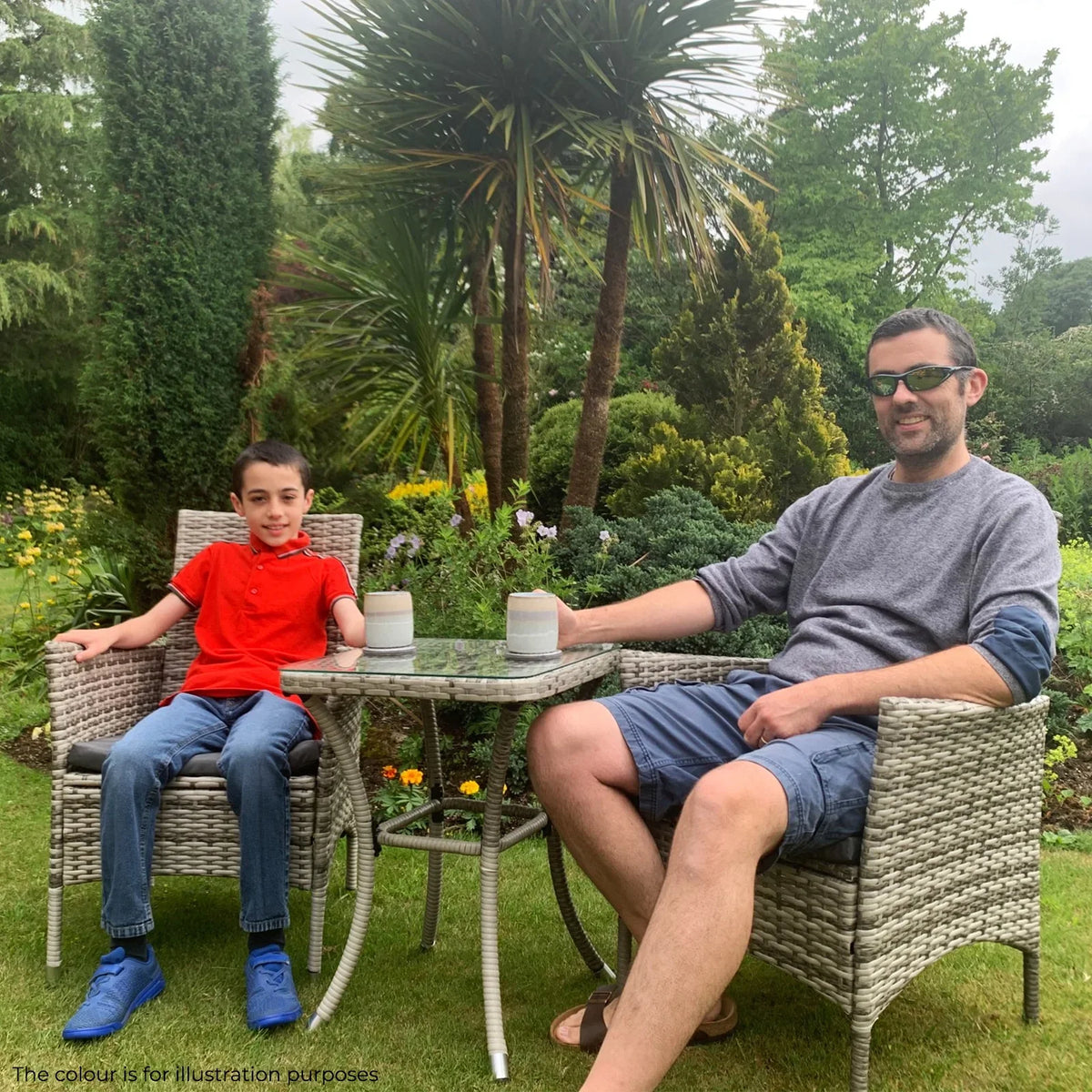 Light grey rattan bistro set with two chairs and a glass-top table, featuring a man and boy enjoying drinks in a garden setting