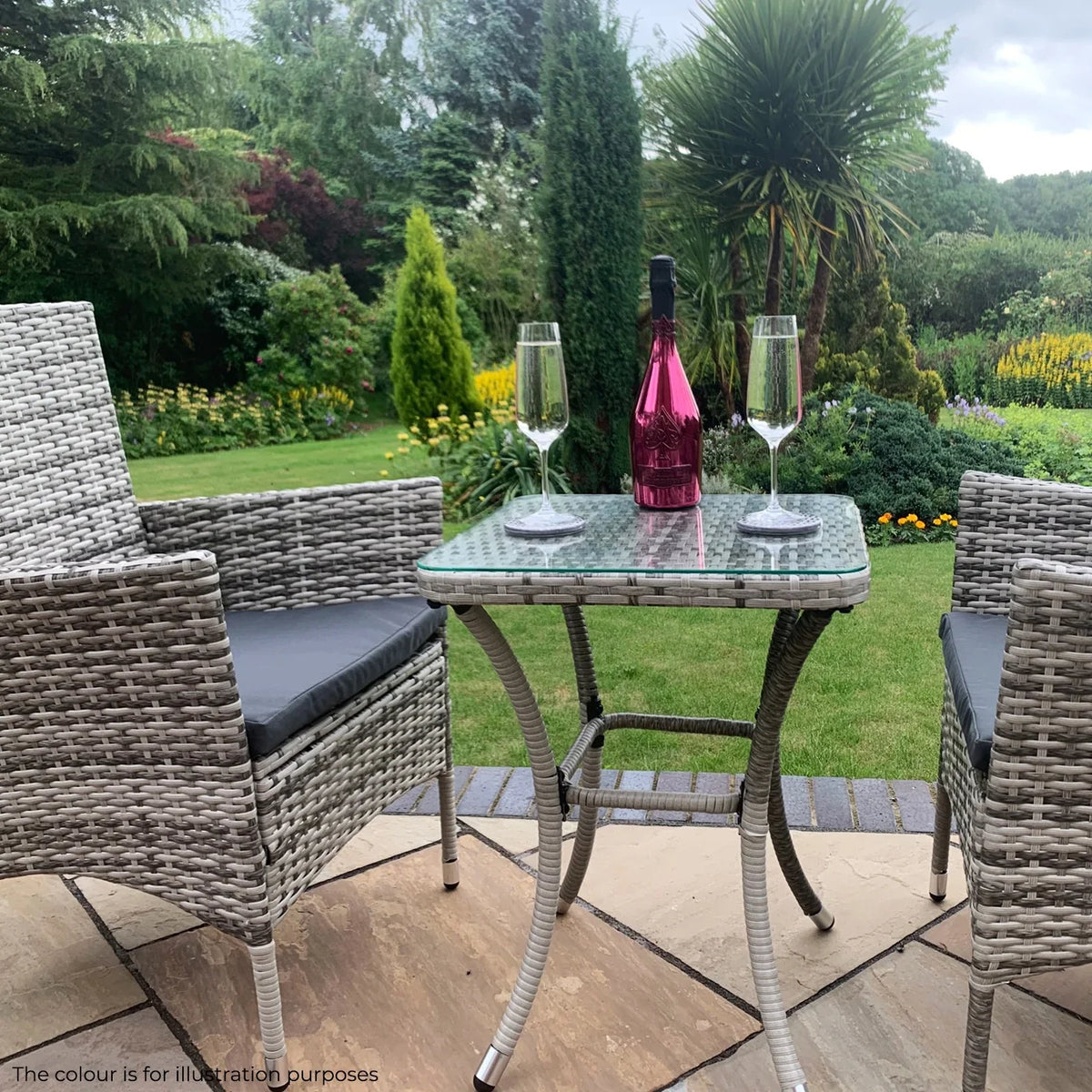 Light grey rattan bistro set with two chairs and a glass-topped table, set in a lush garden with champagne and glasses
