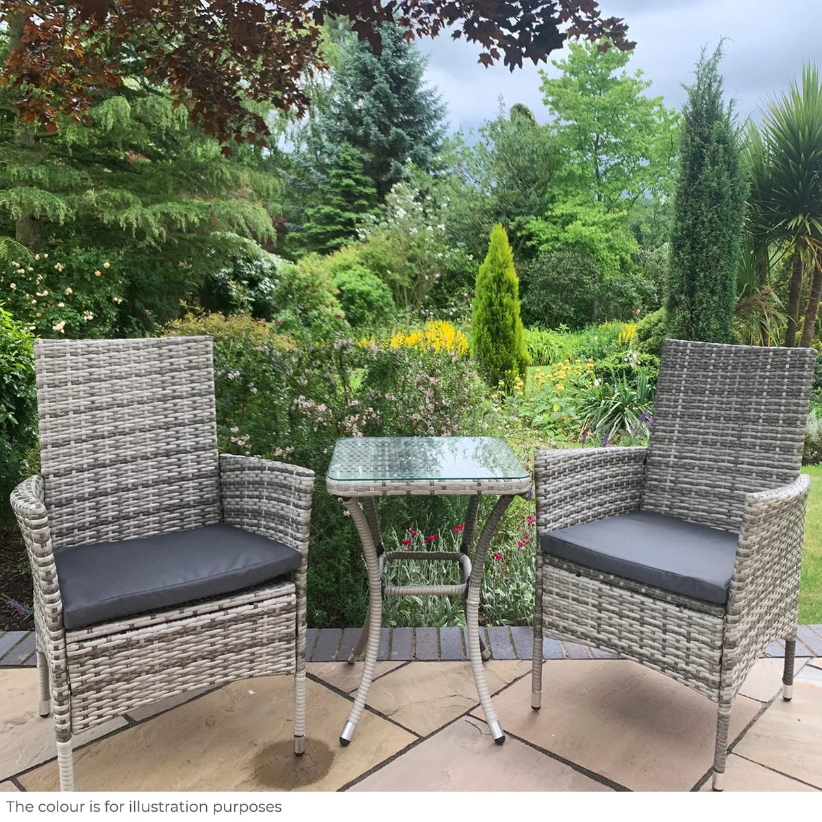 Light grey rattan bistro set featuring two armchairs with cushions and a glass-top table, set against a lush garden backdrop