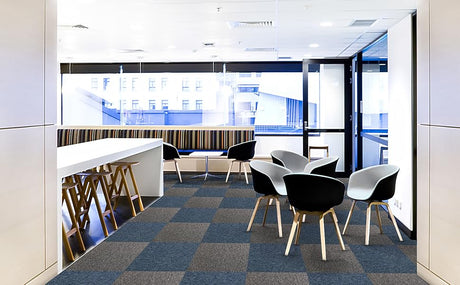 Modern office space featuring gray and blue carpet tiles, sleek white furniture, and stylish black and white chairs with wooden legs