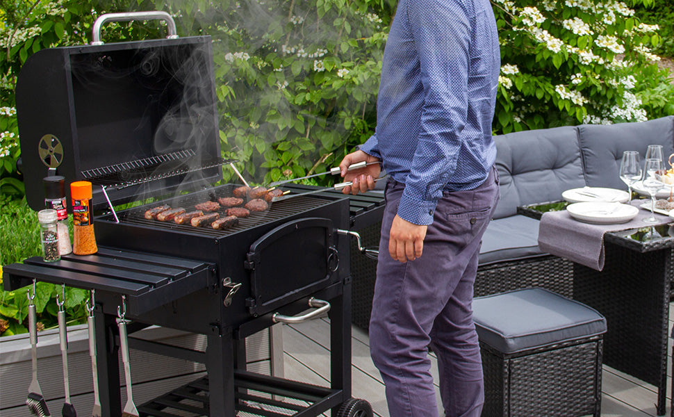 Man grilling sausages on a black gas barbecue, surrounded by greenery and outdoor seating, with smoke rising from the grill
