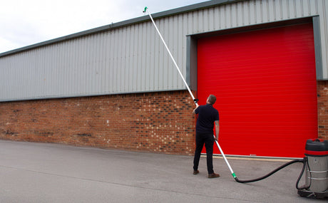 A person using a long pole with a vacuum attachment to clean gutters, standing near a red industrial garage door