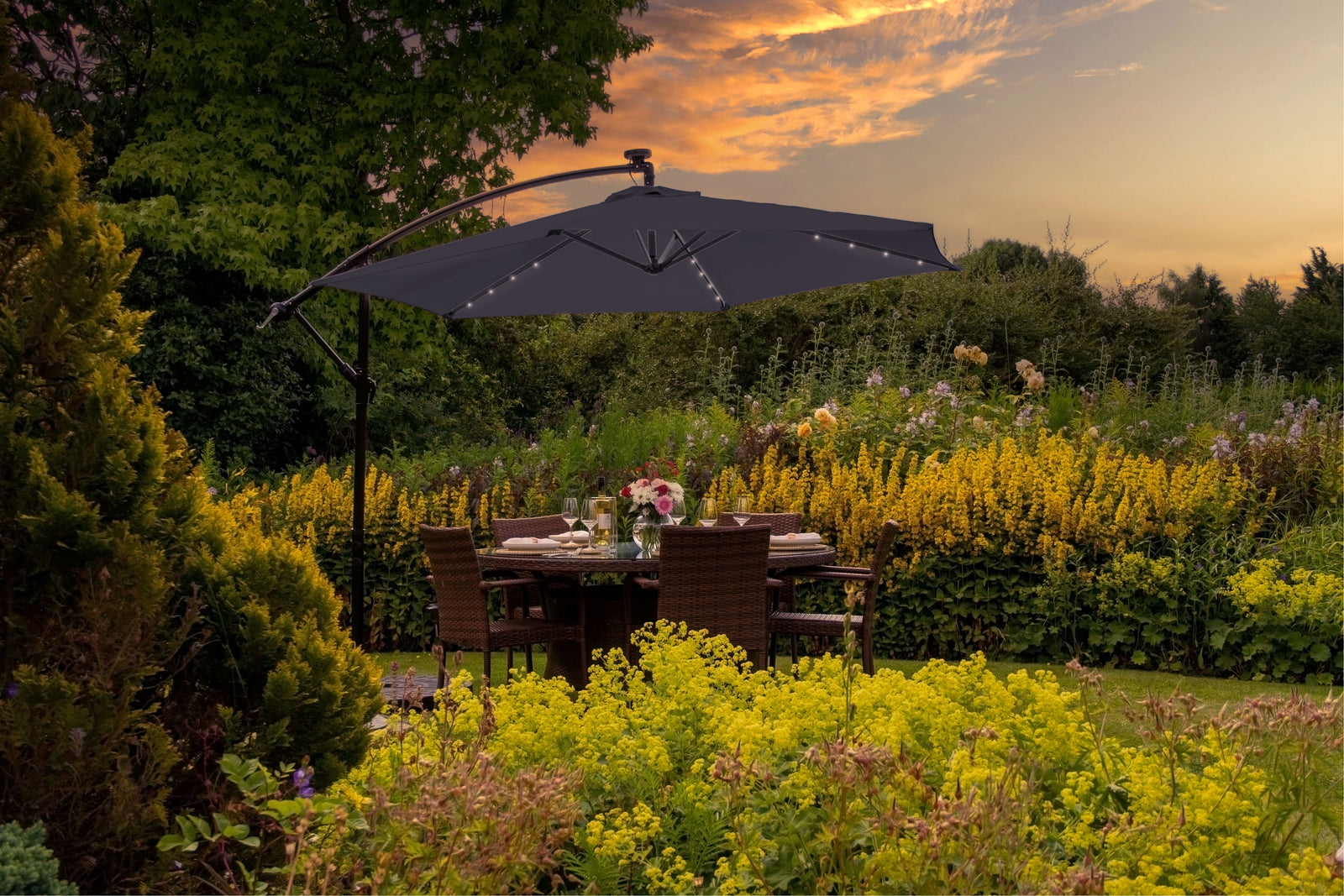 Stylish black garden parasol with LED lights shading a wicker dining set amidst vibrant yellow flowers and lush greenery at sunset
