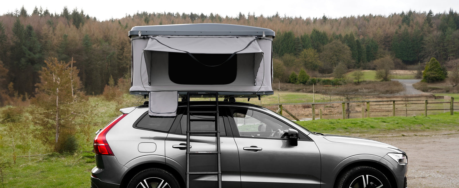 Car roof tent mounted on a silver SUV, featuring a gray canopy, ladder, and scenic forest backdrop, ideal for camping adventures