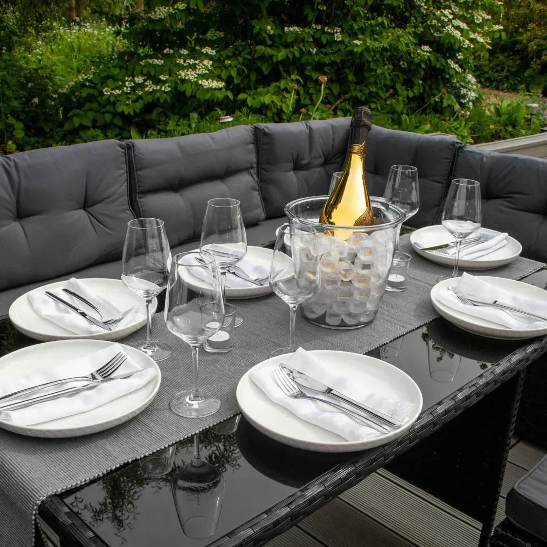 Stylish rattan dining set with gray cushions, elegant glassware, white plates, and a champagne bottle in an ice bucket, set in a lush garden