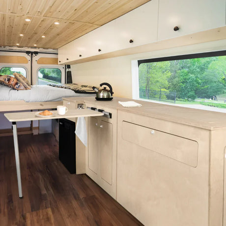Interior of a compact camper van featuring a minimalist wooden design, with a bed at the rear, overhead cabinets, a small kitchenette with a kettle and mini fridge.