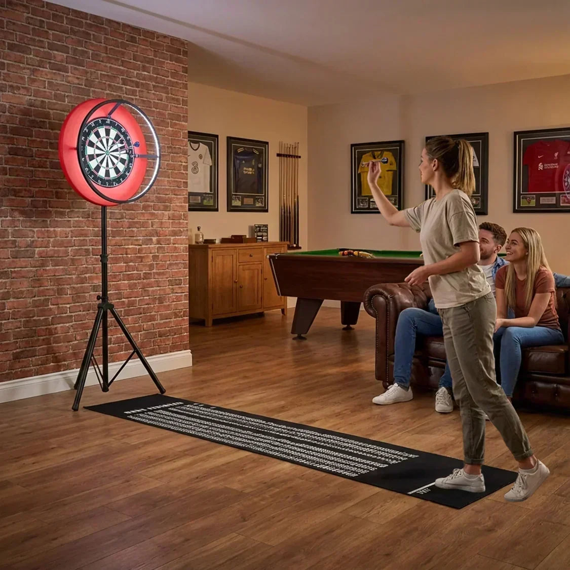 A woman throws a dart at a bright red dartboard, with friends seated on a couch in a cozy home entertainment room