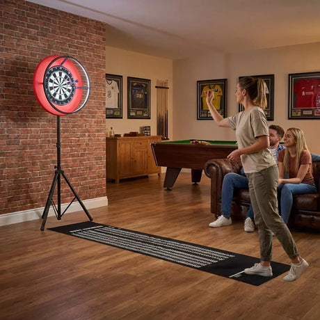 A woman throws a dart at a bright red dartboard, with friends seated on a couch in a cozy home entertainment room