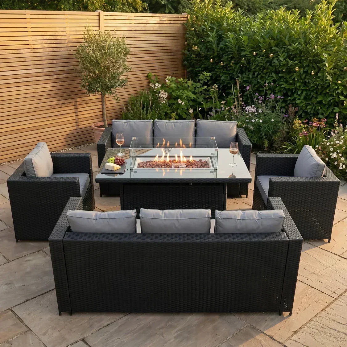 Stylish black rattan garden furniture set with gray cushions, featuring a central fire pit, surrounded by lush greenery and flowers