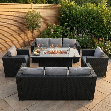 Stylish black rattan garden furniture set with gray cushions, featuring a central fire pit, surrounded by lush greenery and flowers