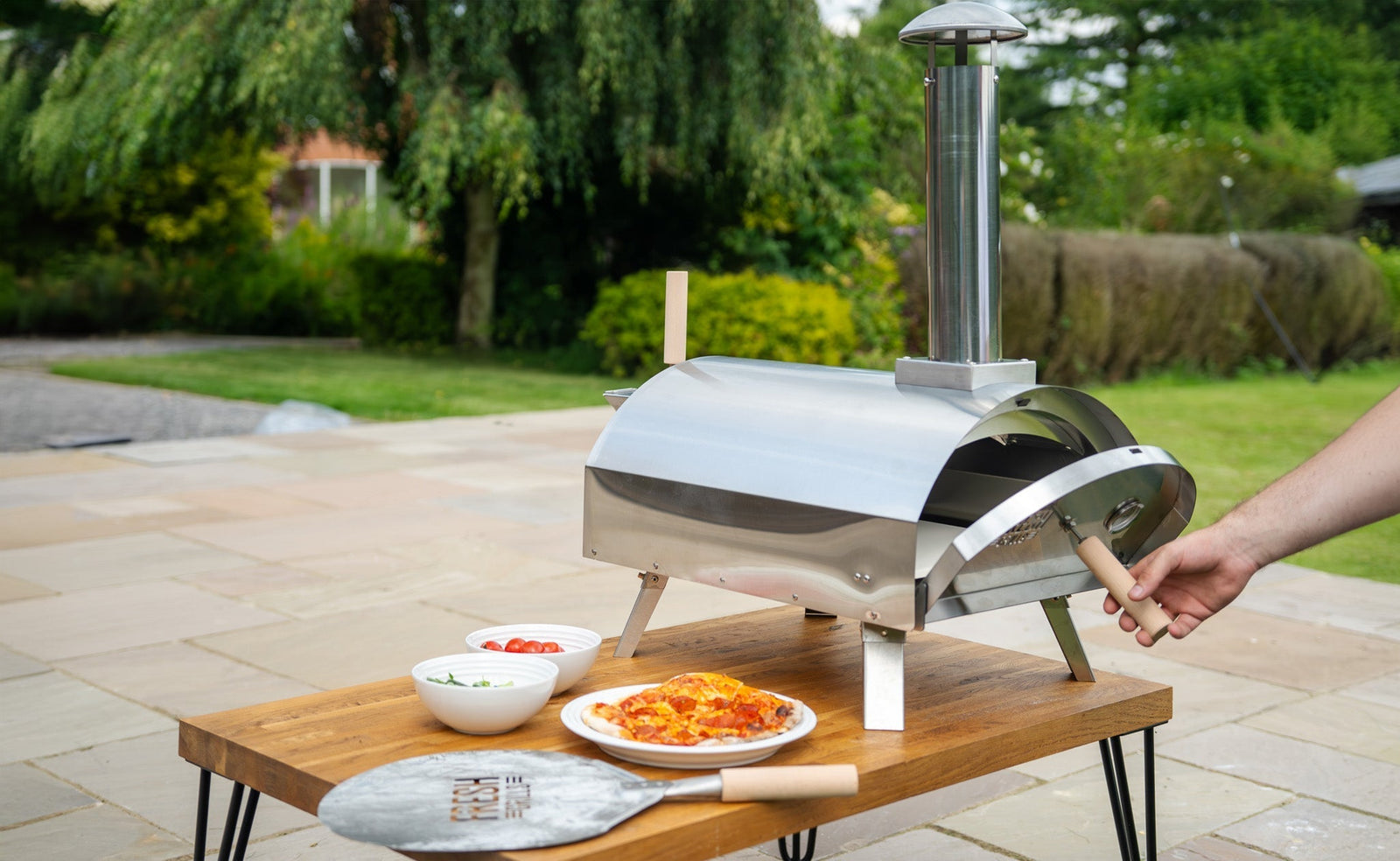Stainless steel pizza oven on a wooden table outdoors, with fresh pizzas and bowls of toppings, surrounded by greenery