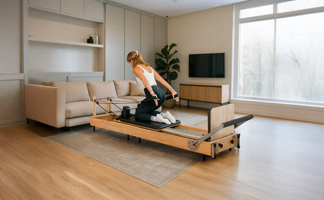 A woman in activewear performs Pilates on a wooden reformer in a bright, modern living room with a sofa and large window