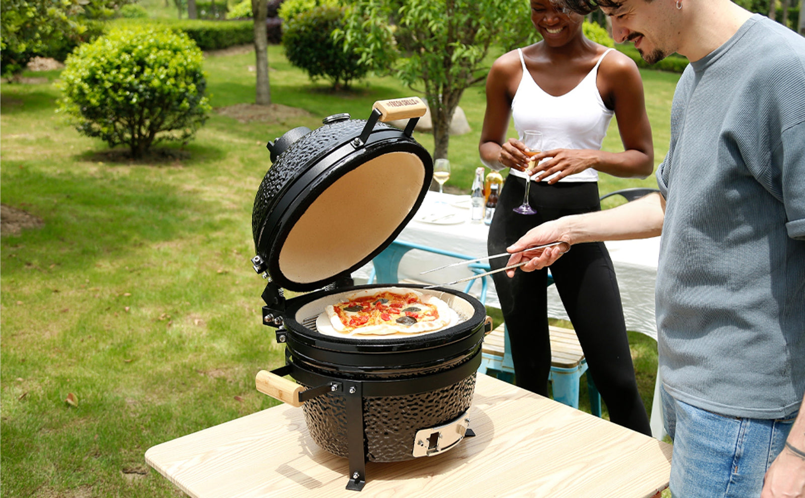 A black ceramic kamado grill with a pizza inside, surrounded by greenery, while a man and woman enjoy drinks nearby