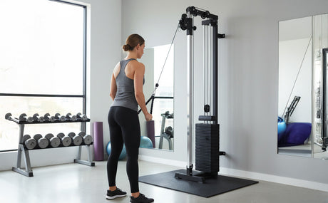 A woman in workout attire uses a wall-mounted cable machine in a modern gym, featuring weights, mirrors, and natural light