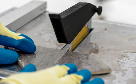 Wet tile cutter in action, with a sharp blade cutting through a gray tile, hands wearing blue gloves guiding the tile carefully
