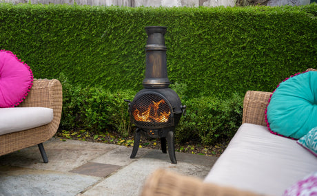A black chiminea with a tall flue, glowing flames inside, surrounded by green hedges and colorful cushions on wicker chairs