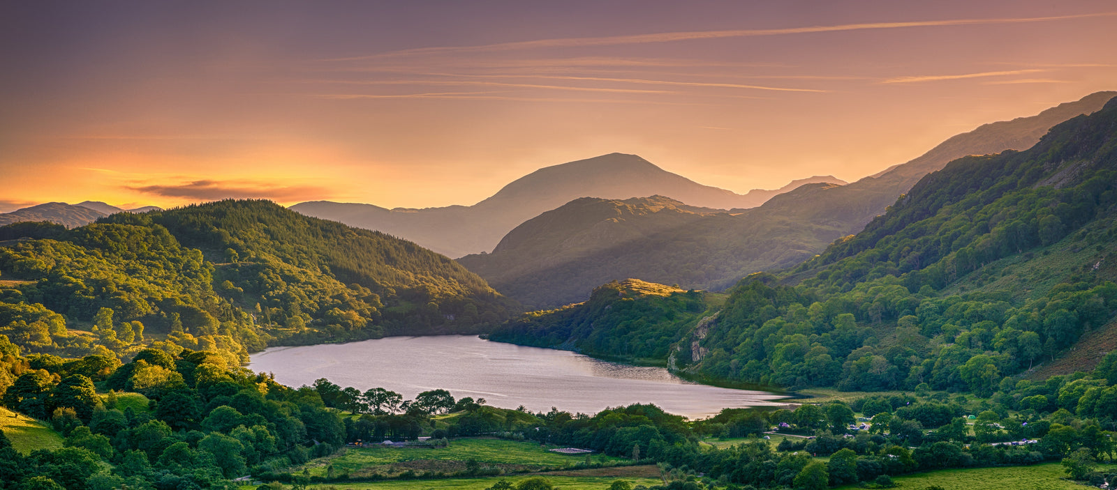 Lush green mountains surround a tranquil lake at sunset, with warm golden light illuminating the serene landscape and sky