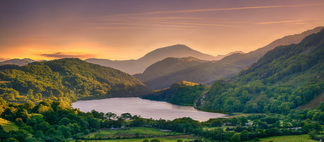 Lush green mountains surround a tranquil lake at sunset, with warm golden light illuminating the serene landscape and sky
