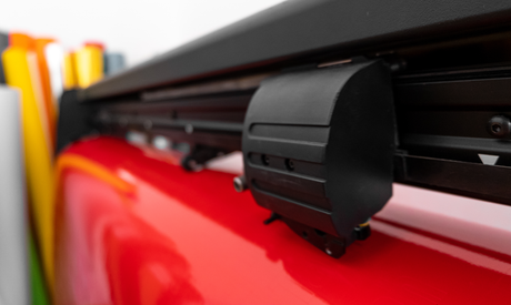 Close-up of a vinyl cutter's black blade holder on a red base, with colorful rolls of vinyl in the background, showcasing its precision and design