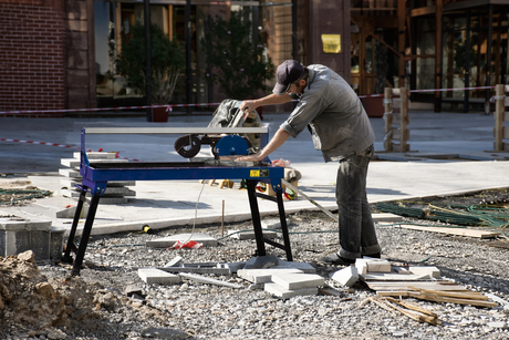 A worker using a wet tile cutter on a construction site, surrounded by gravel, tiles, and tools, wearing a mask and gloves