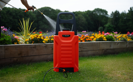 Bright orange garden sprayer positioned on green grass, with colorful flowers in the background and water spraying from a nozzle