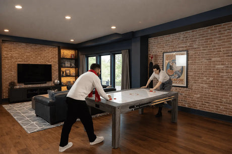 Two people playing air hockey on a sleek, modern 7-in-1 games table in a stylish living room with brick walls and large windows