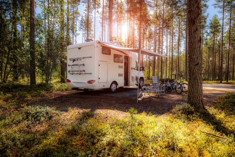 A sleek white motorhome parked in a sunlit forest, surrounded by tall trees, with outdoor chairs and bicycles nearby