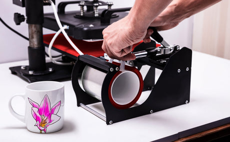A hand adjusts a mug press machine beside a white ceramic mug featuring a vibrant pink orchid design on a clean workspace