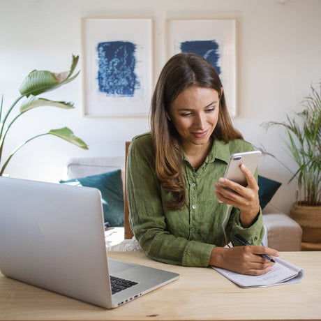 Woman in a green shirt sitting at a wooden desk, smiling at her phone while taking notes, with a laptop and plants in the background