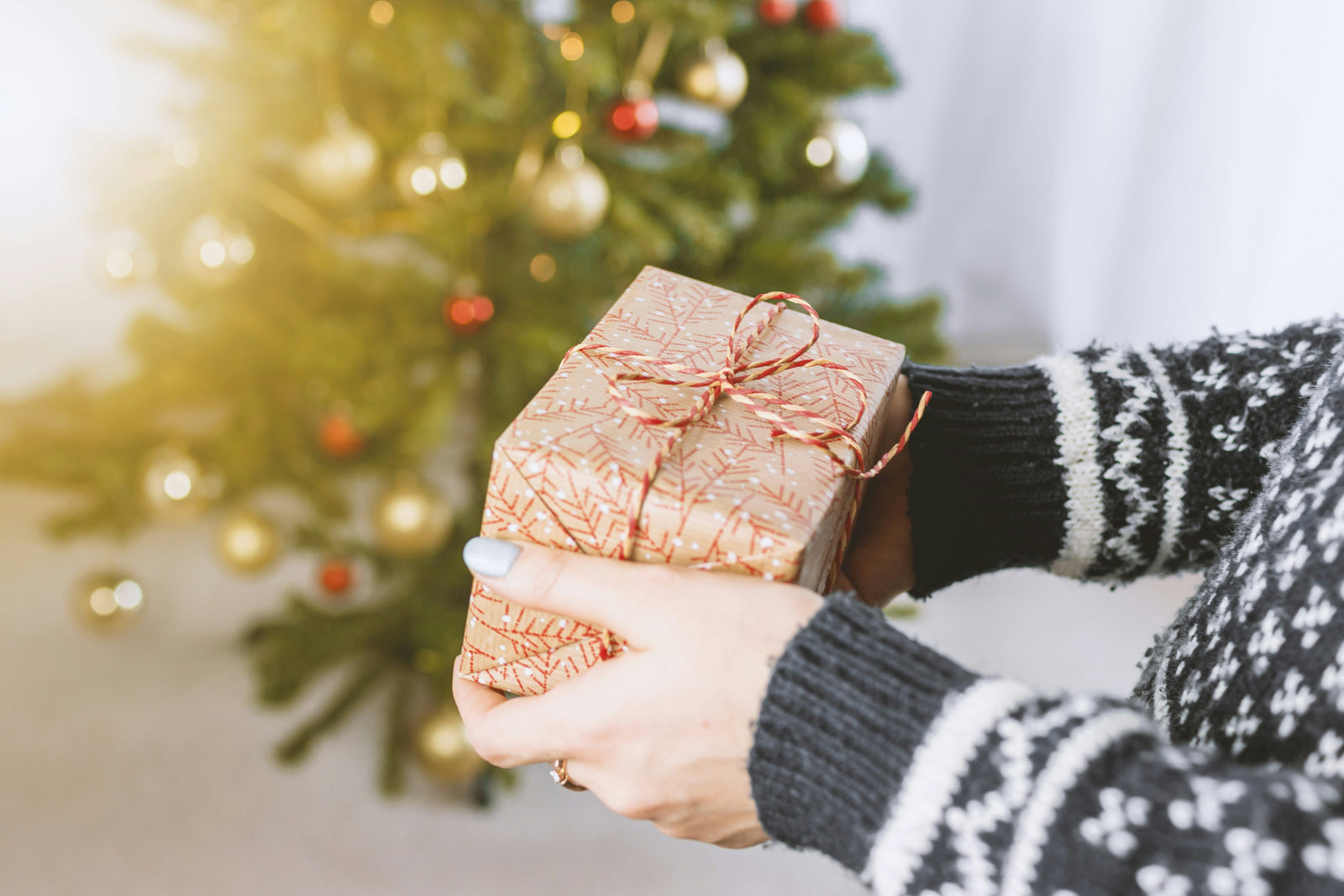 A person in a cozy sweater holds a beautifully wrapped gift with a red bow, standing in front of a decorated Christmas tree