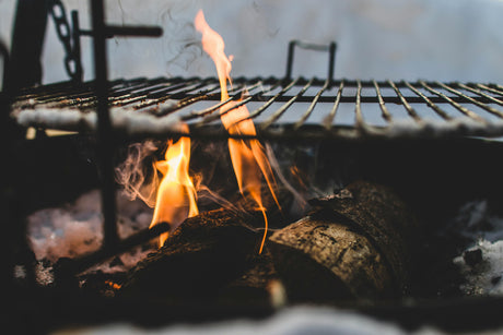 Glowing flames and smoke rise from logs in a grill, showcasing a rustic metal grate perfect for summer BBQ cooking