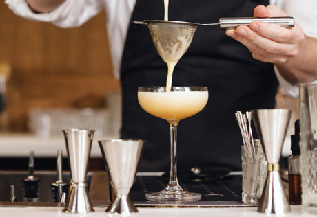 A bartender strains a creamy cocktail into a elegant coupe glass, surrounded by jiggers and bar tools on a polished countertop
