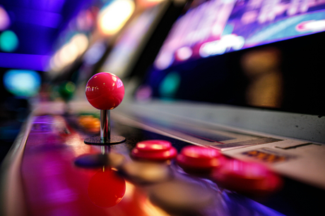 Close-up of a vibrant pink arcade joystick and colorful buttons on a retro arcade machine, set against a blurred neon-lit background