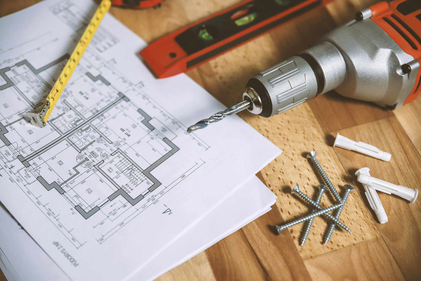 Blueprints of a home renovation project with a drill, screws, and a measuring tape on a wooden surface, showcasing tools for rewiring
