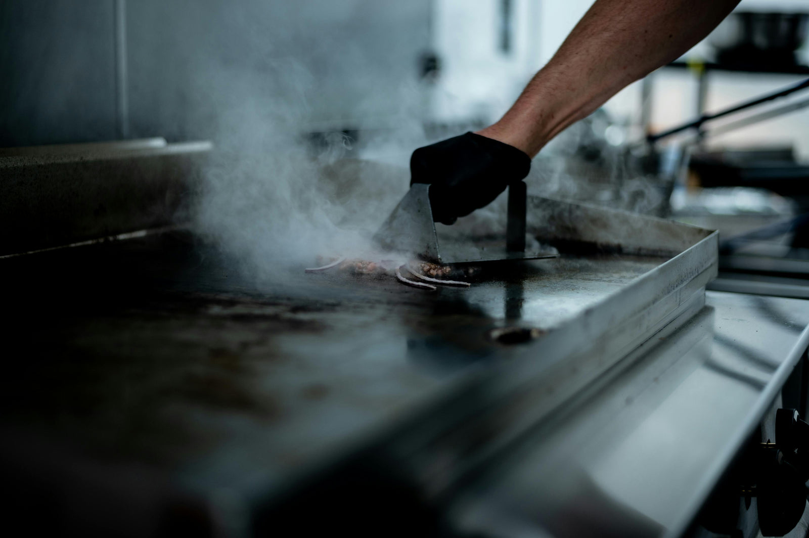 A hand in a black glove flips sizzling food on a shiny electric griddle, surrounded by steam in a modern kitchen setting