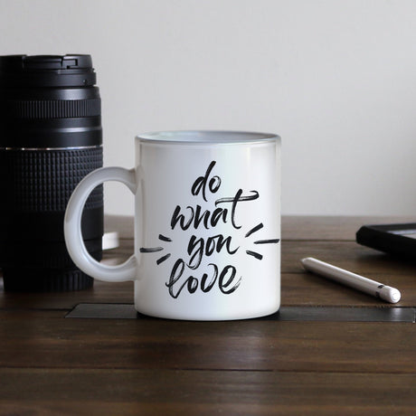 White ceramic mug with black handwritten text "do what you love," placed on a wooden table next to a camera lens and notebook
