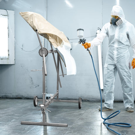 A masked technician in protective gear spray paints a car part on a stand in a well-lit workshop with gray walls and floor