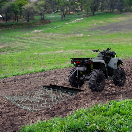 All-terrain vehicle pulling a metal drag harrow across freshly tilled soil in a green, expansive agricultural field