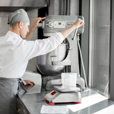 Chef in a white uniform using a silver stand mixer, measuring ingredients in a clear container on a digital scale in a bright kitchen