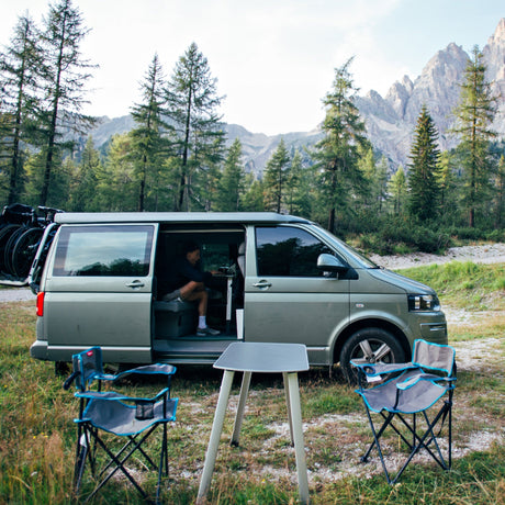 A green motorhome parked in a scenic forest, with a person inside, outdoor chairs, and mountains in the background