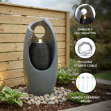Sleek gray electric water feature with ambient LED lighting, cascading water, and a modern design, surrounded by decorative stones and plants