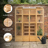 Victorian greenhouse with natural wood finish, glass panels, auto vents, and planter box, set against a brick wall backdrop
