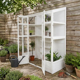 Victorian-style white greenhouse with glass panels, showcasing potted plants, surrounded by greenery and garden tools on stone path